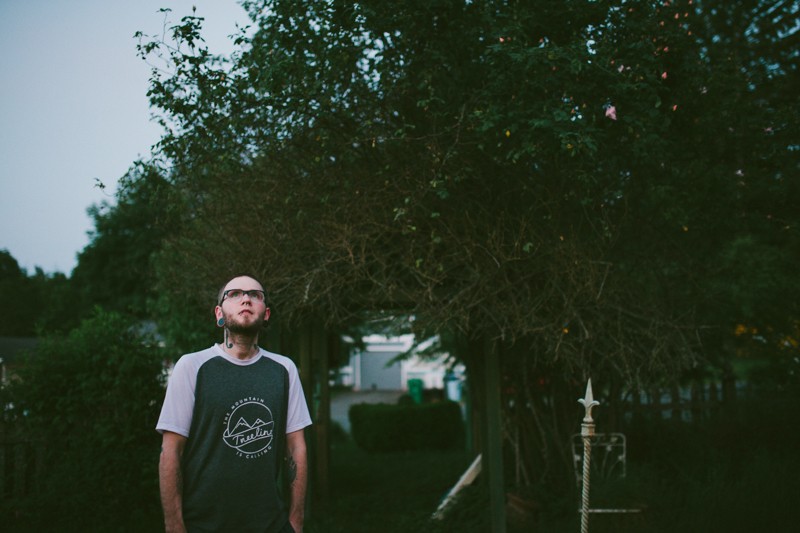 Man in Treeline Outdoors shirt looking up at the moon, with an overgrown Rose bush behind him. 