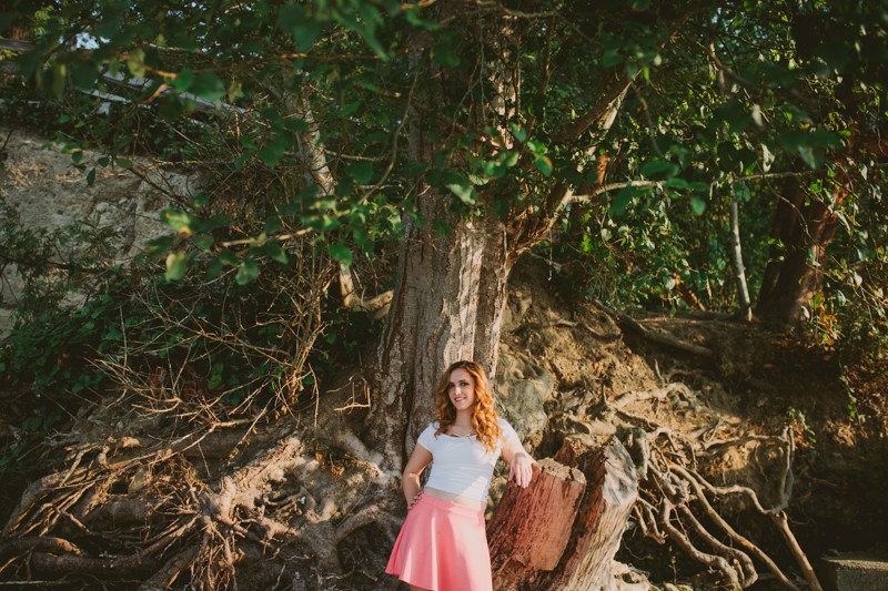 High school senior girl standing in front of a tree, wearing a white shirt and hot pink skirt, at Scenic Beach. 