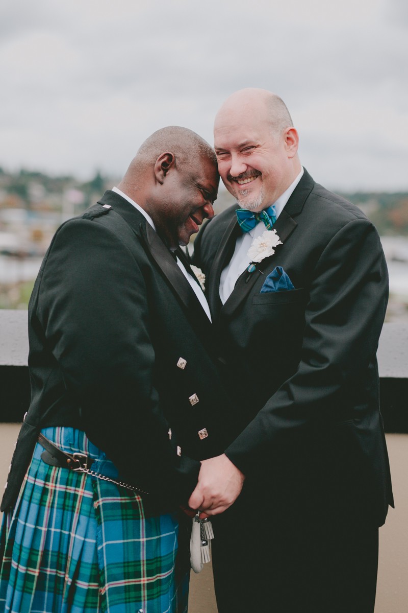 Grooms holding hands and smiling, wearing bow ties and paper flower boutonnieres. 