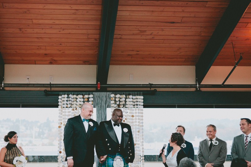 Two grooms holding hands with a paper garland altar backdrop at Olympic Rooftop Pavilion. 