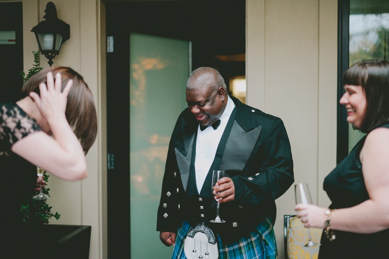 Groom and bridesmaids laughing, with groom in a peak lapel jacket and bow tie, with blue plaid kilt. 