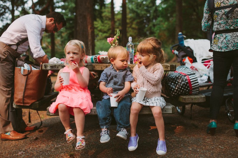Toddlers sitting together snacking on fruit, at a small outdoor wedding. 
