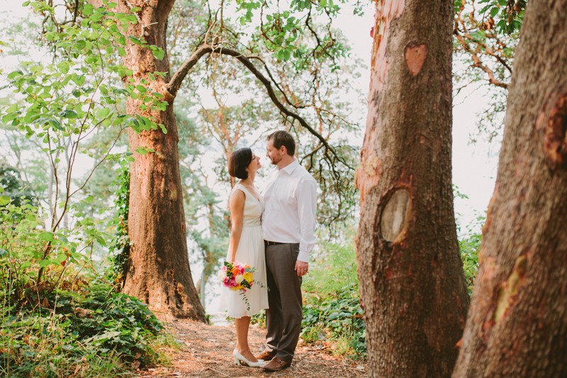 Bride and groom in a forest, hugging, with bride in a simple v-neck dress and groom in a white shirt and slacks. 