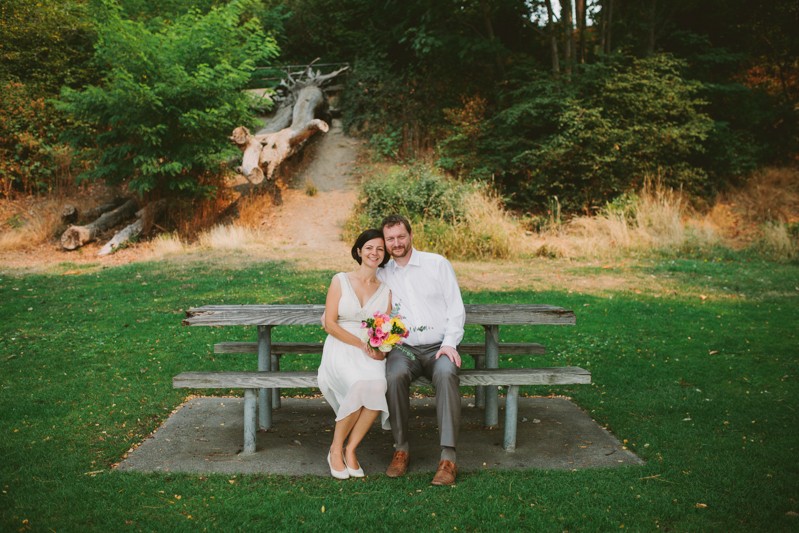 Bride and groom sitting on a picnic table bench, with bride holding a pink and yellow bouquet. 