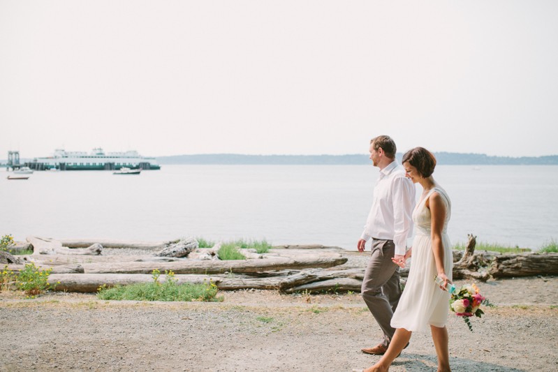 Bride and groom holding hands on the beach, with ferry boat in the distance. 