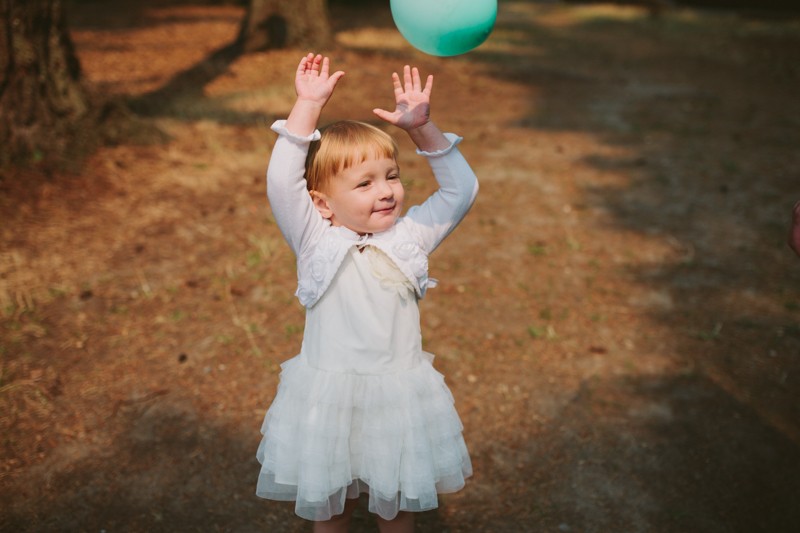 Toddler playing with a balloon at a park wedding, wearing a ruffled white dress and matching cardigan. 