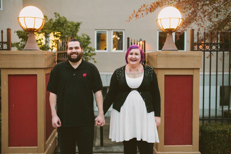 Non-traditional downtown Bremerton engagement session, with alternative bride and groom holding hands in front of lighted columns. 
