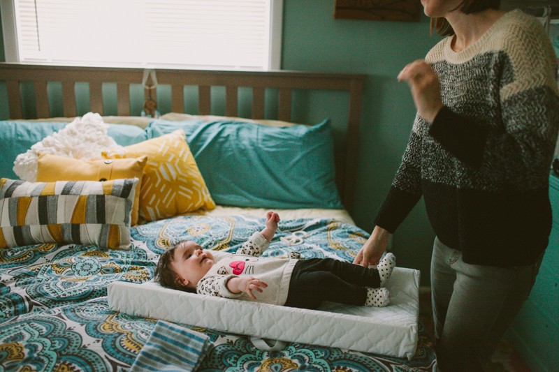 Mom changing baby girl on a bed with a colorful, patterned comforter, in Kitsap County, WA. 
