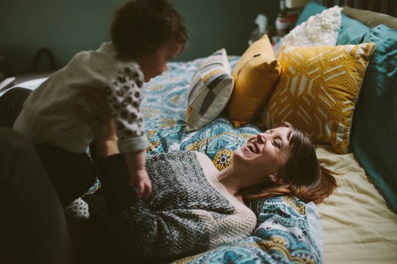 Young mom laying on a colorful bed, laughing, holding her adorable baby. 