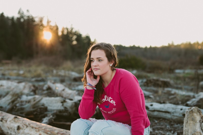 Portraits at the beach, with high school senior girl sitting on driftwood logs at sunset. 