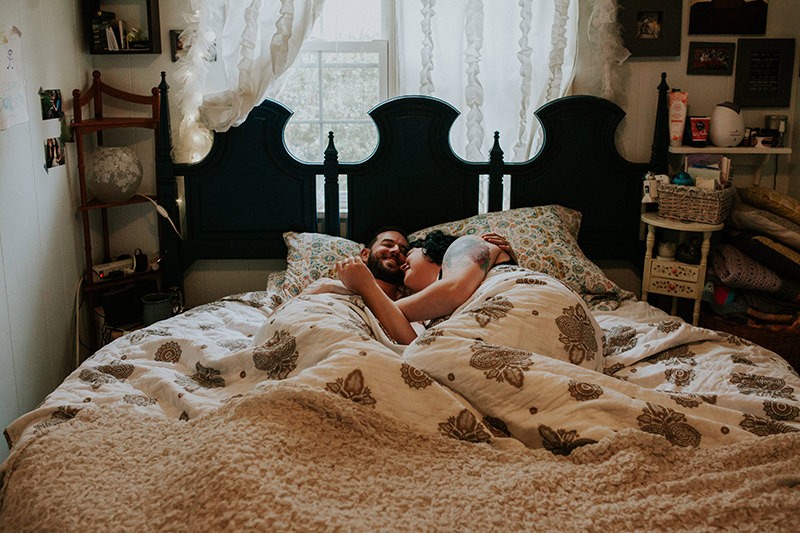 Tattooed indie couple at home, cuddling, with boho bedspread. 