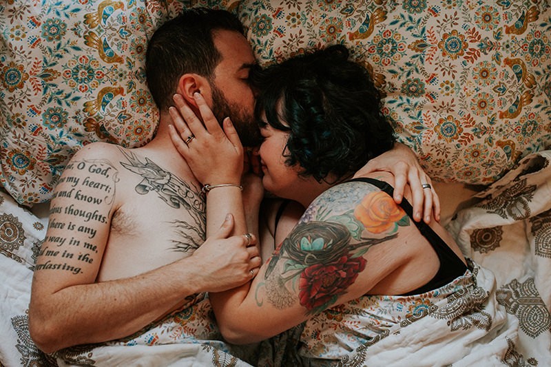 Indie couple cuddling in bed, with tattoos, and boho bed sheets and pillow cases. 