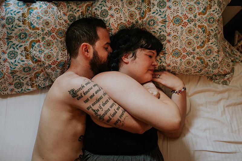 Adorable tattooed couple cuddle on their bed, with patterned boho pillow cases. 