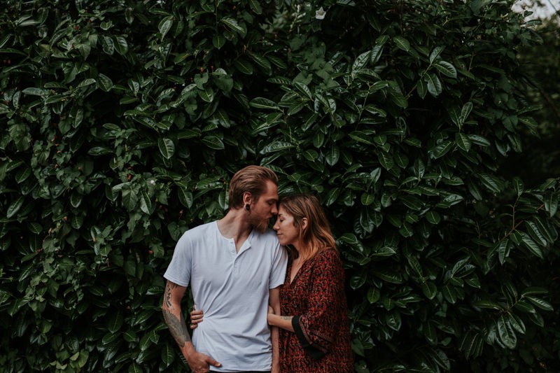 Lifestyle portraits with couple standing in front of a Laurel bush, with woman in a boho dress. 