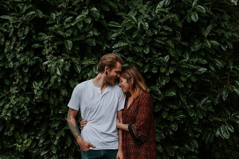 Sweet couple cuddling in front of a Laurel bush, with woman wearing a floral boho dress. 