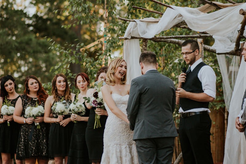 Outdoor garden wedding ceremony, with bride in a strapless beaded dress and bridesmaids in short black dresses. 