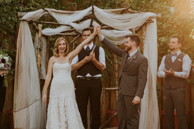 Bride and groom under a rustic wooden altar arch, draped with white fabric. 