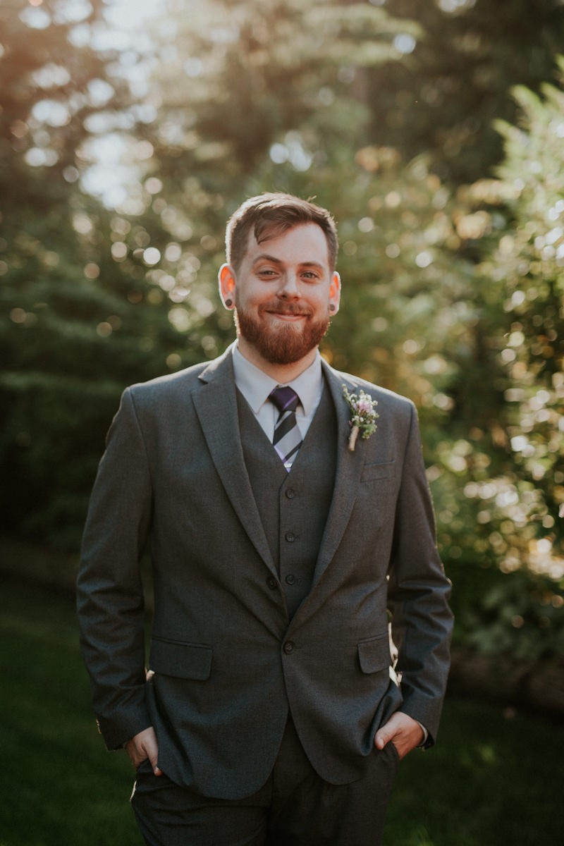 Modern groom wearing grey suit and vest, with a striped purple tie and floral boutonniere. 