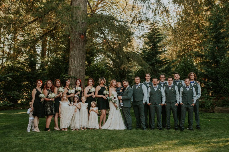 Large wedding party, with bridesmaids in black dresses and groomsmen in grey vests and bow ties. 