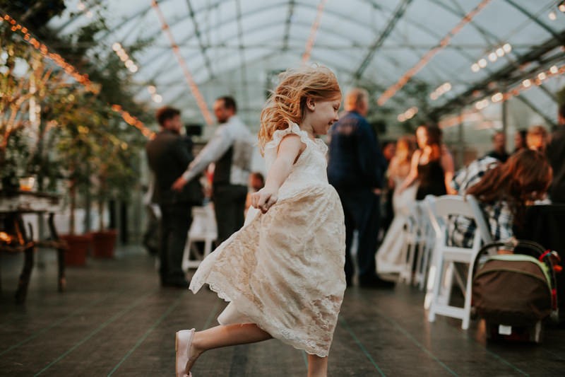 Darling flower girl in white lace dress, dancing in a greenhouse venue at sunset. 