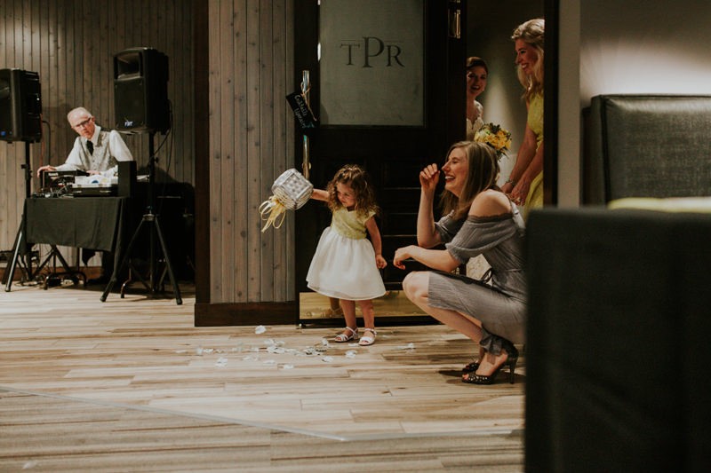 Flower girl dumps petals out of her basket as bridesmaids and bride laugh. 