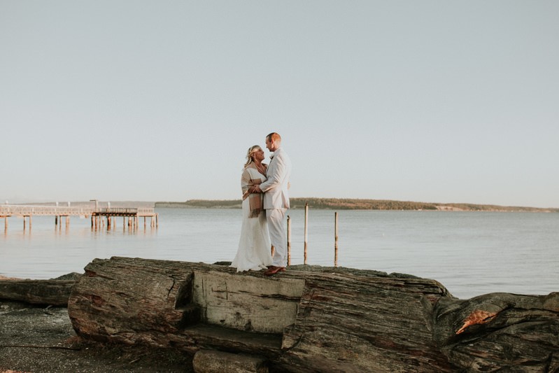 Bride and groom stand on a large log on the Port Townsend waterfront. 