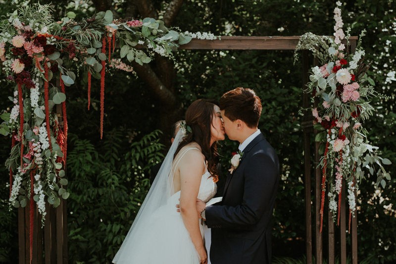 Modern bride and groom kiss under wooden arch with floral details. 